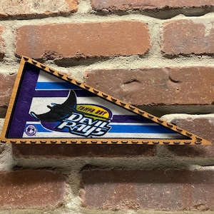 Puede incluir: Un banderín triangular de madera con un logotipo de los Tampa Bay Devil Rays. El logotipo presenta una pelota de béisbol azul y amarilla con las palabras "Tampa Bay Devil Rays" en ella. Una raya manta estilizada está detrás de la pelota. El banderín tiene un fondo a rayas azul y blanco.