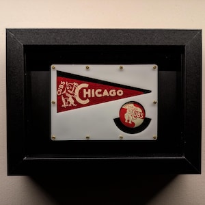Puede incluir: Bandera y botón enmarcados de los Chicago Cubs. La bandera es roja y blanca con la palabra "Chicago" y el logotipo de los Cubs. El botón presenta el logotipo de los Cubs. El marco es negro.
