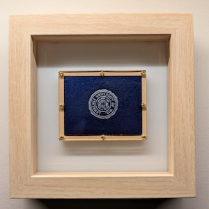 May include: A framed blue fabric patch with gold tacks and the Lawrence University seal in white.