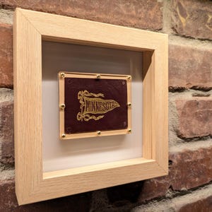 May include: A framed pennant with the word "Minnesota" in gold lettering on a maroon background. The pennant is set within a light wood frame with a white mount. Small gold tacks secure the pennant to the frame.