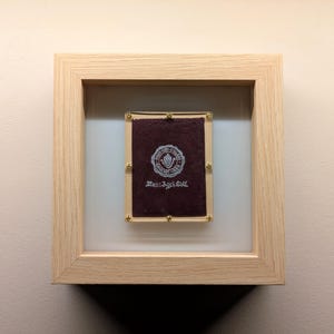 May include: A framed piece of paper with a red background and a white seal with the text "Mag-Az-Sol" in the centre. The frame is made of light wood.