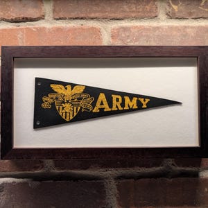 May include: Framed black pennant with gold lettering and emblem that reads "ARMY". The pennant is in a dark brown wooden frame with a white mount. The background is a brick wall.