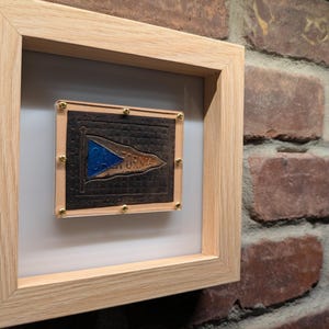 May include: Framed artwork with a brown plaque displaying a blue flag design and the word "CALIFORNIA". The plaque is set against a white backdrop within a light wood frame. Gold studs secure the plaque to the frame.