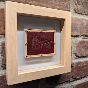 May include: A framed piece featuring a burgundy leather square with the word "KANSAS" embossed on it. The leather square is secured with gold-colored studs and set against a white background within a light wood frame.