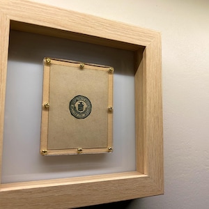 Op de afbeelding: Een ingelijst stuk papier met een groene zegel en gouden punaises. De zegel heeft een ontwerp van een gebouw met een vlag erop.