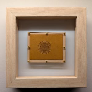 Könnte beinhalten: Ein gerahmtes Kunstwerk mit braunem Lederhintergrund und einem goldenen Siegel mit dem Text "The University of Alabama" in der Mitte.