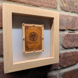 May include: A framed piece featuring a brown leather item with the text "Princeton Univ" and a university emblem. The item is secured with gold-coloured studs and set against a white background within a light wood frame. The frame is mounted on a brick wall.