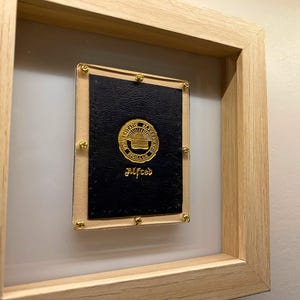 May include: A framed black leather-bound book with gold accents and the text "Alfred" on the cover. The book is displayed in a wooden frame with a white mat.