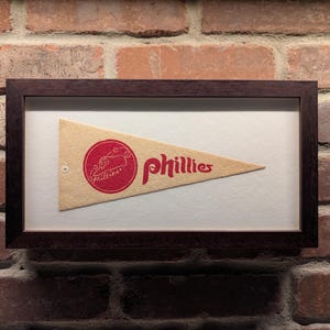 May include: Framed vintage Phillies pennant. The pennant is beige with red lettering and a circular logo. The frame is dark brown and the background is white. The pennant is displayed against a brick wall.