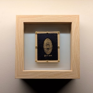 May include: A framed piece of art with a gold design on a black background. The frame is made of light wood and has a glass front.