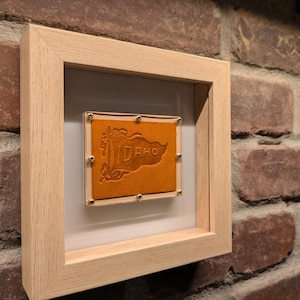 May include: A framed artwork featuring a tan leather piece with the word "IDAHO" embossed on it. The leather is secured with brass-colored tacks within a light wood frame, set against a white background. The frame is mounted on a brick wall.