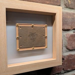 May include: A framed piece of brown leather with gold-coloured rivets and an embossed design. The leather is in a light wood frame.