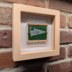 Puede incluir: Un banderín de Dartmouth enmarcado con fondo verde y texto blanco. El banderín está montado en un marco de madera clara con un paspartú blanco. El marco está montado en una pared de ladrillo.