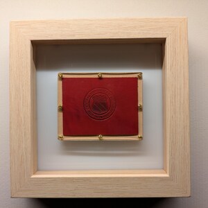 May include: A framed red leather patch with gold accents and a crest featuring a shield and the text "The National Hockey League" in a white circle.