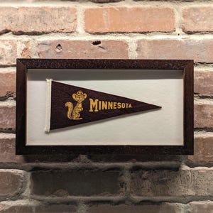 Könnte beinhalten: Gerahmtes Vintage-Wimpel mit braunem Hintergrund, mit einem goldenen Eichhörnchen-Maskottchen und dem Wort "MINNESOTA" in goldener Schrift. Der Wimpel wird in einem dunkelbraunen Holzrahmen vor einer Backsteinmauer ausgestellt.
