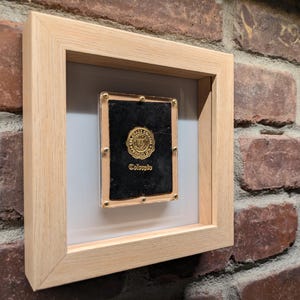 May include: A framed gold medallion, inscribed with "Colorado", is presented. The medallion is set against a black backdrop within a light-coloured wooden frame. The frame is mounted on a brick wall.