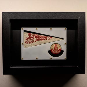 May include: A framed pennant featuring a red and white Washington Senators logo and a red, white and blue Washington Nationals button. The pennant is displayed in a black frame with gold accents.