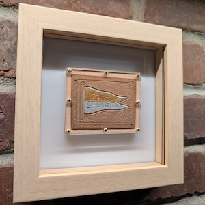 Può includere: Un'opera d'arte da parete quadrata incorniciata con un design a gagliardetto. Il gagliardetto è marrone chiaro con le parole "LEHIGH" e "LEMON" in azzurro. La cornice è in legno chiaro, fissata con piccole viti dorate.