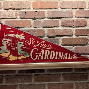 May include: A red pennant displaying "St. Louis Cardinals" in white script. The pennant features a cartoon cardinal bird with a baseball bat. The background is a brick wall.