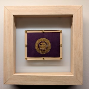 May include: A framed image of a gold seal with a purple background. The seal features a book, a dove, and the text "Universitas Thomensis 1843".
