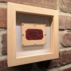 May include: A framed piece of art featuring a red, textured stamp with the word "DENVER" embossed on a tan background. The artwork is encased in a light wood frame and mounted on a brick wall.