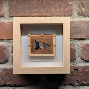 Könnte beinhalten: Ein gerahmtes Kunstwerk mit einer braunen Lederflagge mit goldenen Akzenten und einem schwarz-weißen Design. Die Flagge ist auf einem weißen Hintergrund montiert und in einem hellen Holzrahmen gerahmt.