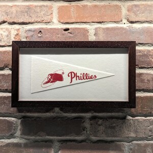 May include: Framed pennant with the Philadelphia Phillies logo. The white pennant has a red Phillies logo and text. The dark brown frame contrasts against the brick wall background.