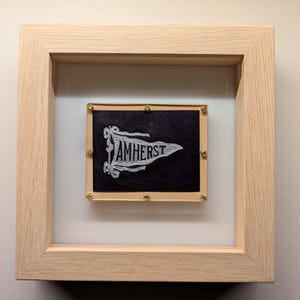 Könnte beinhalten: Ein gerahmtes schwarz-weißes Wimpel mit dem Wort "AMHERST" in weiß gestickt. Der Wimpel wird in einem hellen Holzrahmen mit goldenen Nägeln präsentiert.
