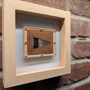 May include: A framed leather patch with the text "USMC" and a stylised eagle design. The patch is mounted on a white background and has gold-coloured tacks around the edges.