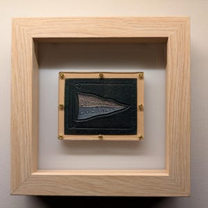 May include: A framed artwork featuring a black and gray pennant with the text "FOLLOW YOUR DREAMS" in white. The pennant is displayed in a light wood frame with gold accents.