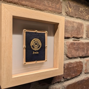 May include: A framed emblem featuring the word "Duke" below a gold emblem on a navy blue background. The emblem is in a gold frame, mounted in a light wood frame. The frame is mounted on a brick wall.