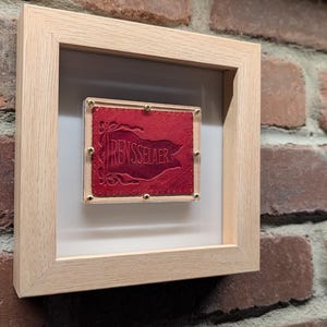 May include: A framed artwork featuring a red leather piece with the word "RENSSELAER" embossed on it. The artwork is encased in a light wood frame with a white mat, mounted on a brick wall.