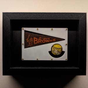 Könnte beinhalten: Eine schwarze Schattenbox mit weißem Hintergrund. In der Schattenbox befindet sich ein Vintage-Wimpel der St. Louis Browns und ein Baseball mit dem Logo der St. Louis Browns.