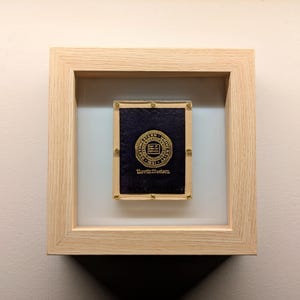 May include: A framed piece featuring a navy blue plaque with a gold seal and the text "Northwestern". The plaque is mounted within a light-colored wooden frame, creating a shadow box effect. The frame is square and has a natural wood grain.