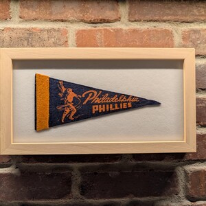 May include: Framed Philadelphia Phillies pennant. The pennant is navy blue with orange accents and features the team name and a baseball player graphic. The pennant is displayed in a light wood frame against a brick wall.