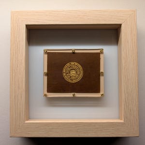 May include: A framed image of a gold seal with the text "Founded 1865 The University of Chicago" surrounded by a laurel wreath. The seal is on a brown background and is framed in a light wood frame.