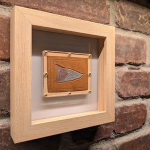 May include: A framed artwork displaying a brown leather pennant with the word "CHICAGO" and a flag design in silver. The pennant is set against a light grey backdrop within a wooden frame, held by brass-coloured screws.