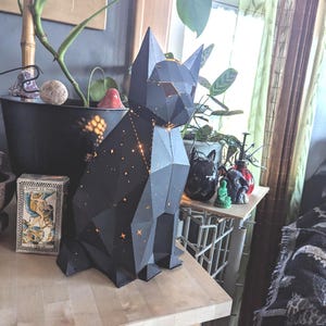 May include: A black geometric cat-shaped night light with a constellation design. The light is illuminated and casting a warm glow.