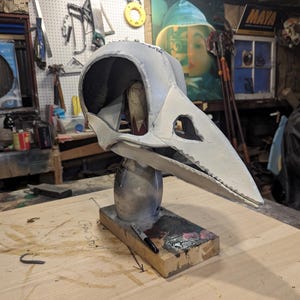 May include: A grey and white raven skull mask, mounted on a silver-painted stand and a wooden base. The mask has a large beak and eye holes. A black marker and paint are on the wooden base. The background is a workshop.