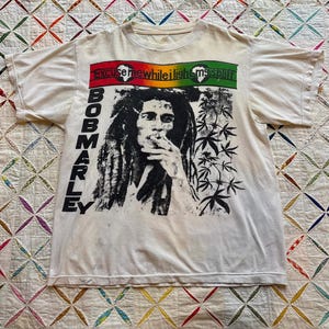 Peut inclure: T-shirt crème avec un portrait en noir et blanc de Bob Marley. Le t-shirt comprend le texte "BOB MARLEY" verticalement sur le côté gauche et la phrase "Excuse me while I light my spliff" en haut en rouge, jaune et vert.