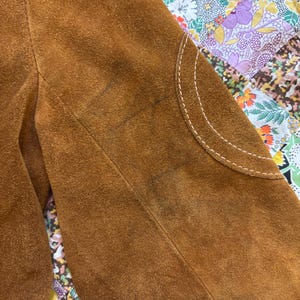 Puede incluir: Primer plano de una manga de chaqueta de ante marr&oacute;n. La chaqueta presenta un parche curvo con costuras blancas. El ante tiene un aspecto texturizado con algunas manchas m&aacute;s oscuras. El fondo es un estampado floral colorido.