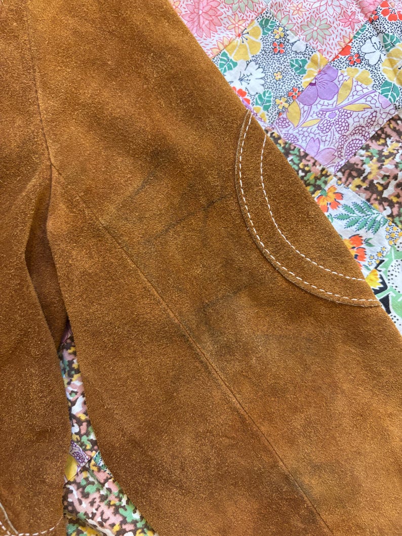 Puede incluir: Primer plano de una manga de chaqueta de ante marr&oacute;n. La chaqueta presenta un parche curvo con costuras blancas. El ante tiene un aspecto texturizado con algunas manchas m&aacute;s oscuras. El fondo es un estampado floral colorido.