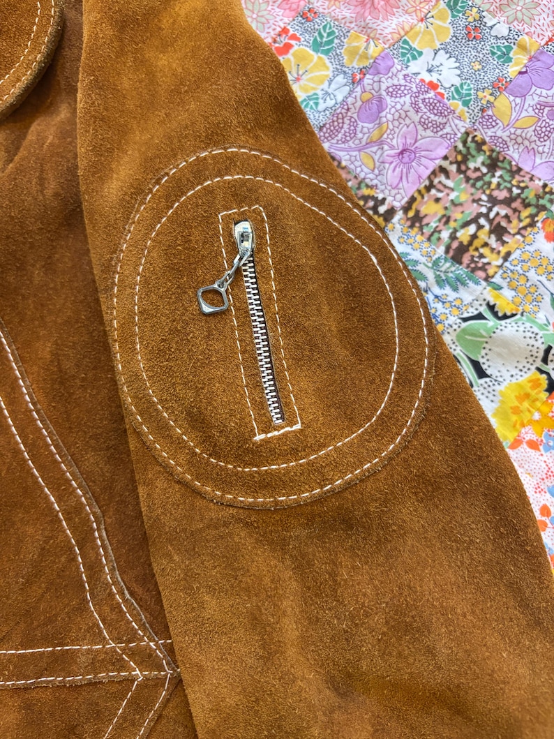 Puede incluir: Primer plano de una chaqueta de ante marr&oacute;n con costuras blancas y una cremallera plateada. La chaqueta presenta un parche circular con un bolsillo con cremallera. El ante tiene un aspecto suave y texturizado. El fondo es un estampado floral colorido.