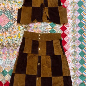 May include: A two-piece set featuring a vest and a skirt. The vest and skirt are made of brown and dark brown suede, with a patchwork design. The skirt has a button-up front. The set is displayed on a colorful quilt.