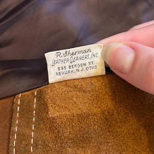 Puede incluir: Primer plano de un art&iacute;culo de cuero de gamuza marr&oacute;n con costuras blancas. Una etiqueta con el texto "R. Sherman LEATHER CLEANERS, INC. 895 BERGEN ST. NEWARK, N.J. 07112" es visible.