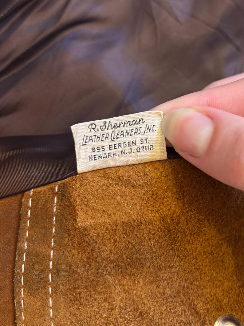 Puede incluir: Primer plano de un art&iacute;culo de cuero de gamuza marr&oacute;n con costuras blancas. Una etiqueta con el texto "R. Sherman LEATHER CLEANERS, INC. 895 BERGEN ST. NEWARK, N.J. 07112" es visible.