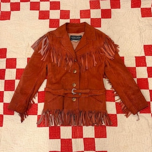 Puede incluir: Chaqueta de ante color óxido con flecos en las mangas, el pecho y el dobladillo. La chaqueta tiene un cinturón con hebilla plateada y cierres de botón. La etiqueta dice "COSA NOVA".