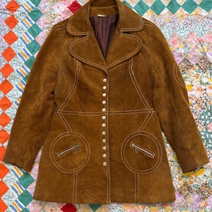 Puede incluir: Una chaqueta vintage de ante marr&oacute;n con cuello de solapa, cierre de botones y dos bolsillos delanteros con cremalleras. La chaqueta presenta detalles de costura blancos y un forro interior marr&oacute;n. La chaqueta se muestra sobre un edred&oacute;n patchwork colorido.