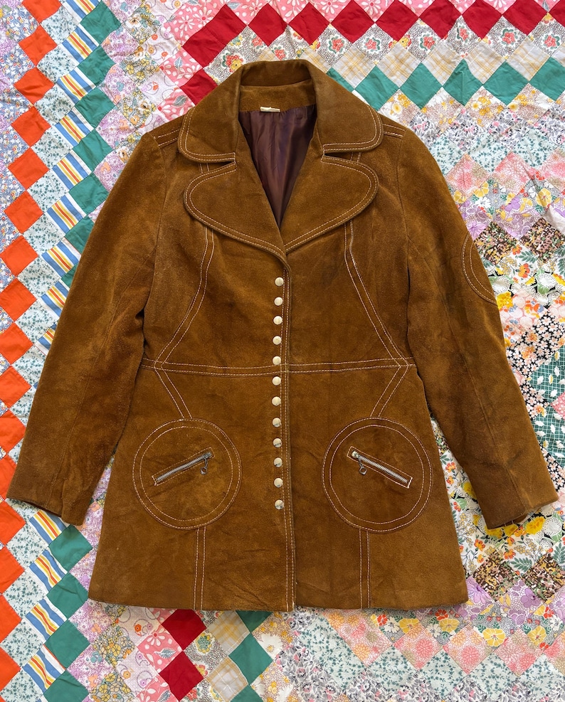 Puede incluir: Una chaqueta vintage de ante marr&oacute;n con cuello de solapa, cierre de botones y dos bolsillos delanteros con cremalleras. La chaqueta presenta detalles de costura blancos y un forro interior marr&oacute;n. La chaqueta se muestra sobre un edred&oacute;n patchwork colorido.