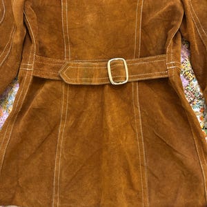 Puede incluir: Chaqueta de ante marr&oacute;n con cintur&oacute;n a juego y hebilla dorada. La chaqueta presenta costuras blancas a lo largo de las costuras y un corte entallado. La chaqueta es de estilo vintage.